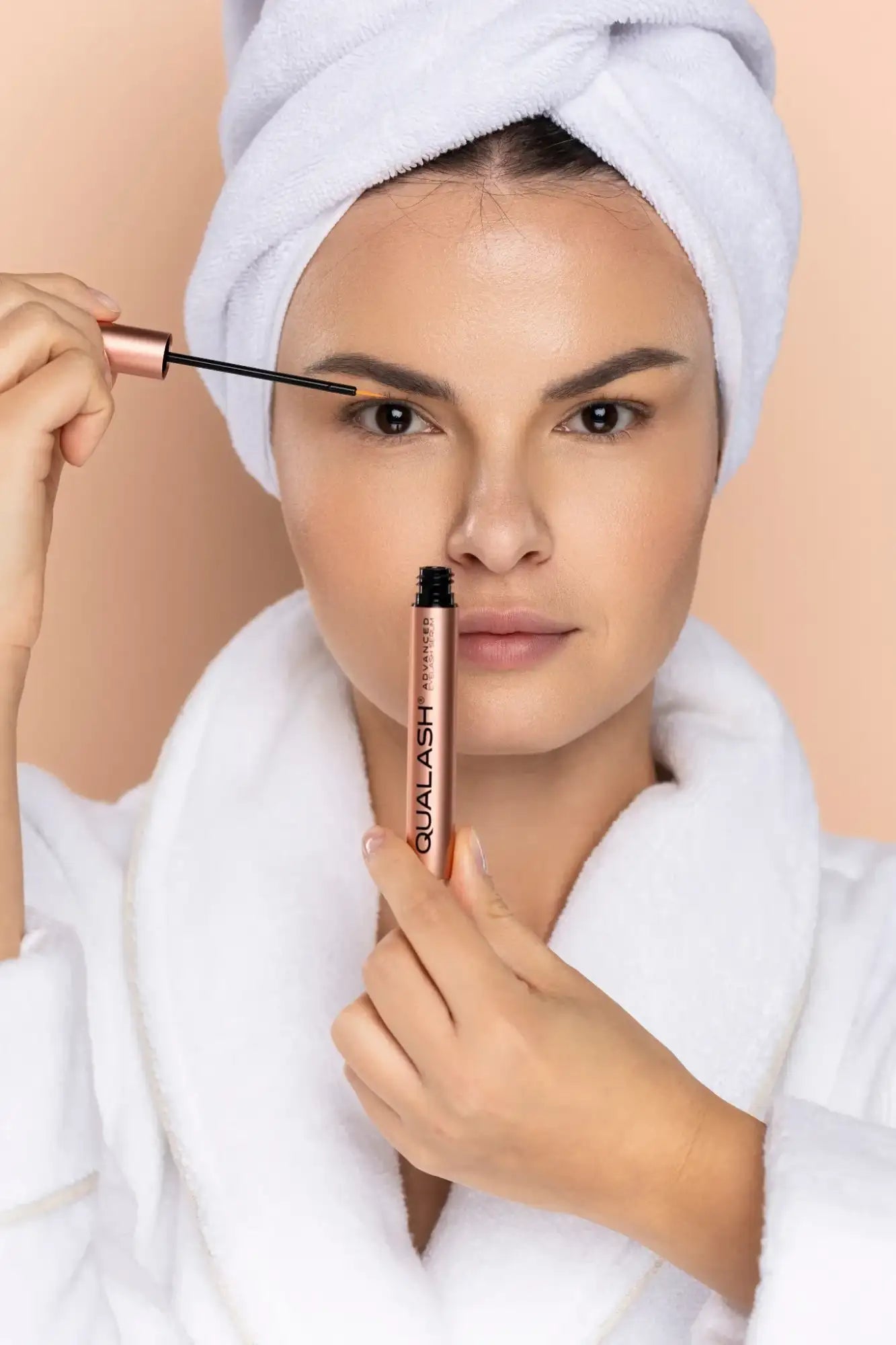 Front-facing beauty portrait of a woman holding a Qualash cosmetic tube and applying brow serum with an applicator on a neutral background